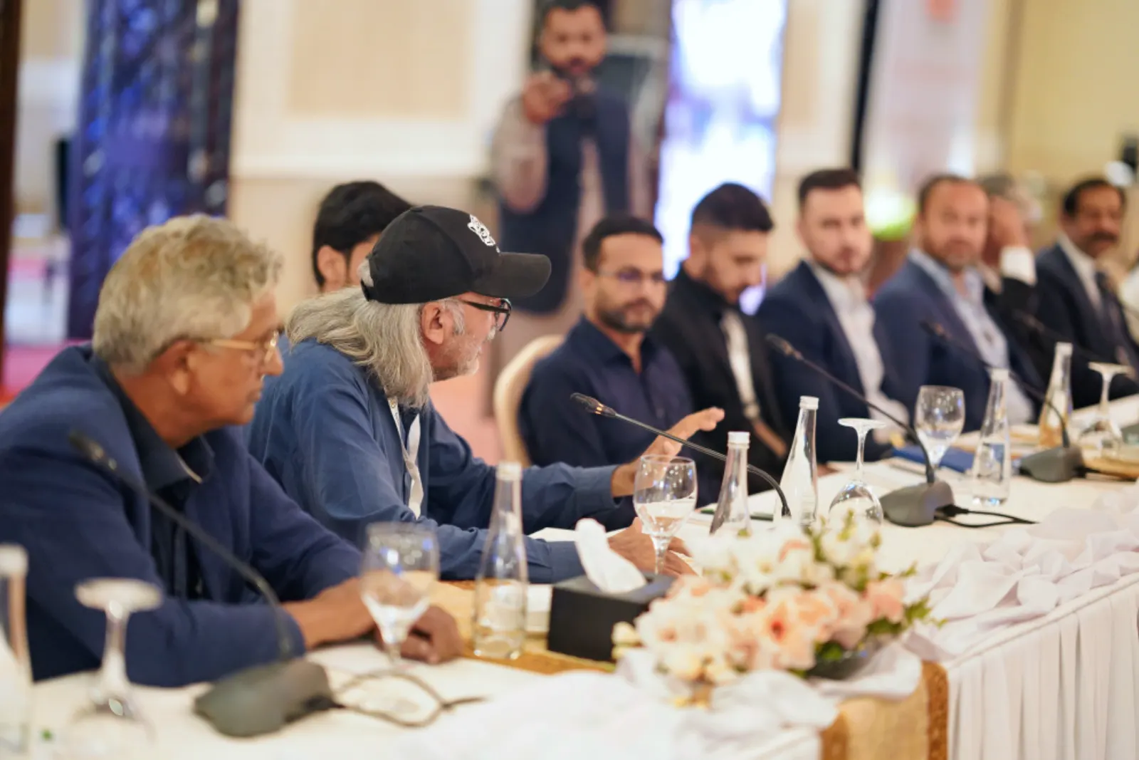 A delegate in a cap makes a point to the delegates seated along the conference table.