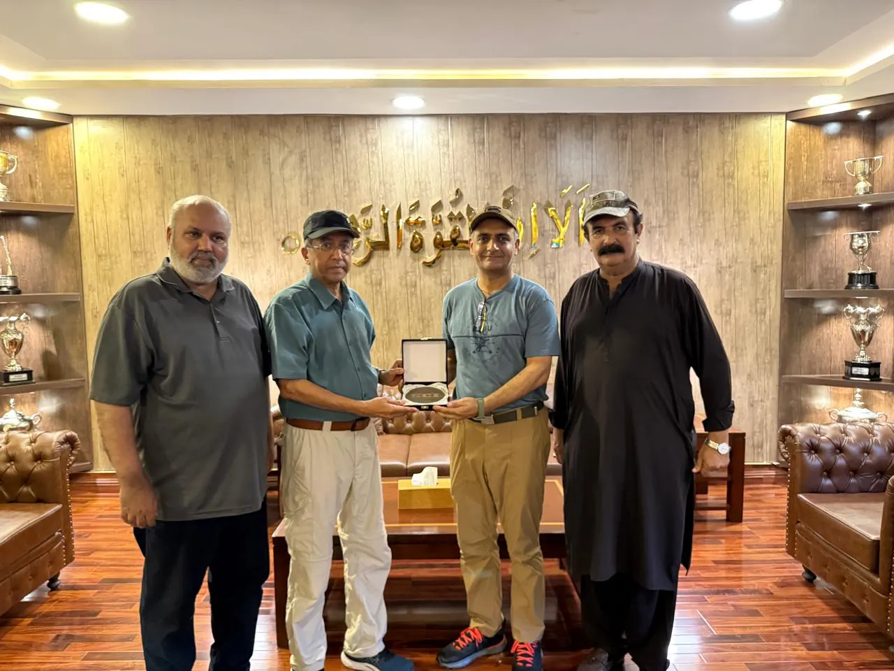 Four chapter members hold a presentation plaque in a trophy room lined with cups.