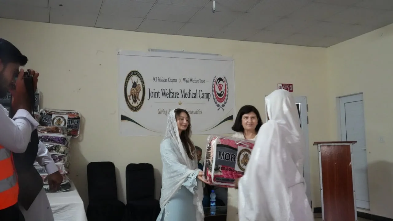 A woman in a pale blue dress hands a blanket to a woman in white beside the camp banner.