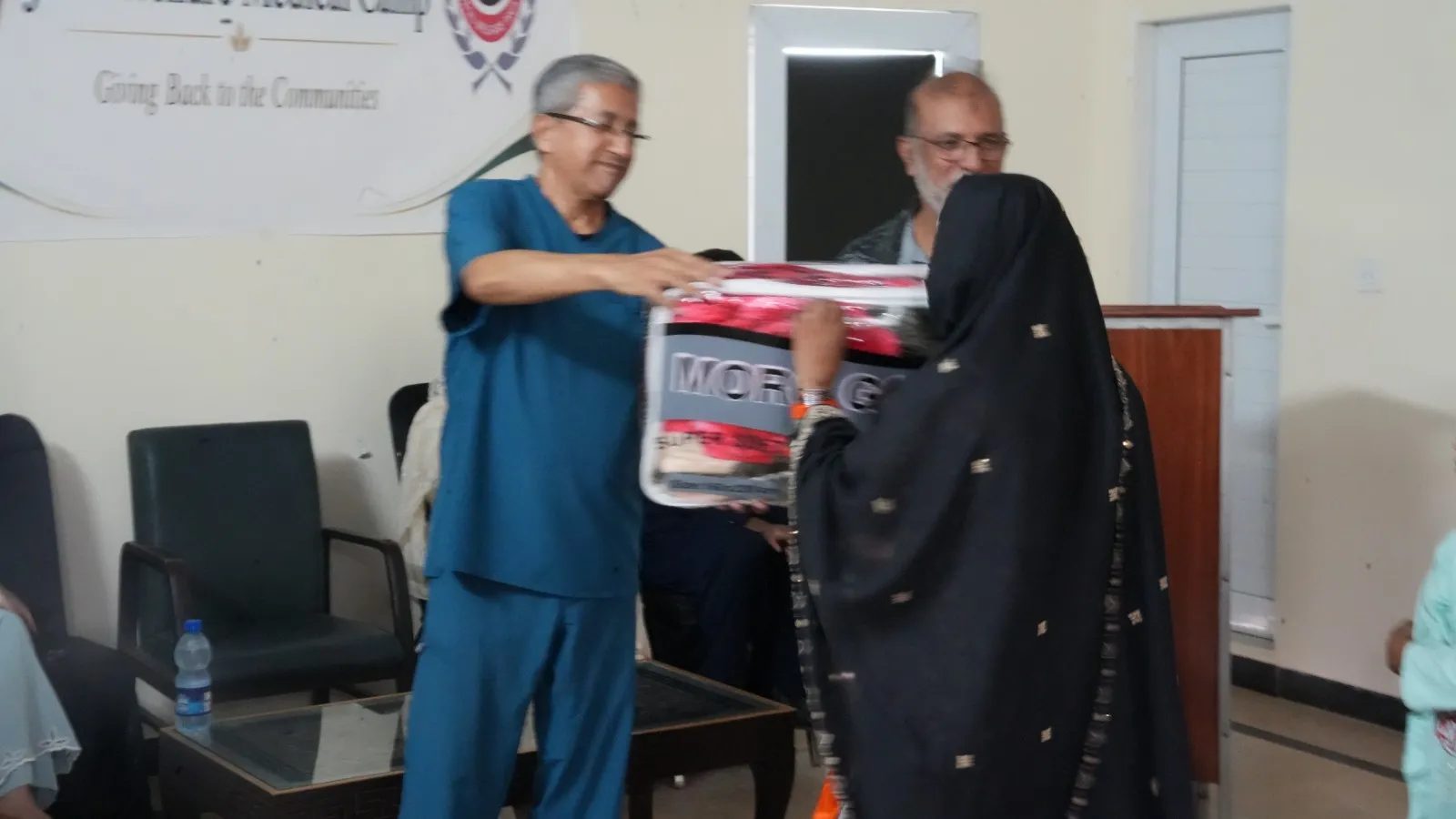 A doctor in blue scrubs hands a wrapped blanket to a woman beside the lectern.