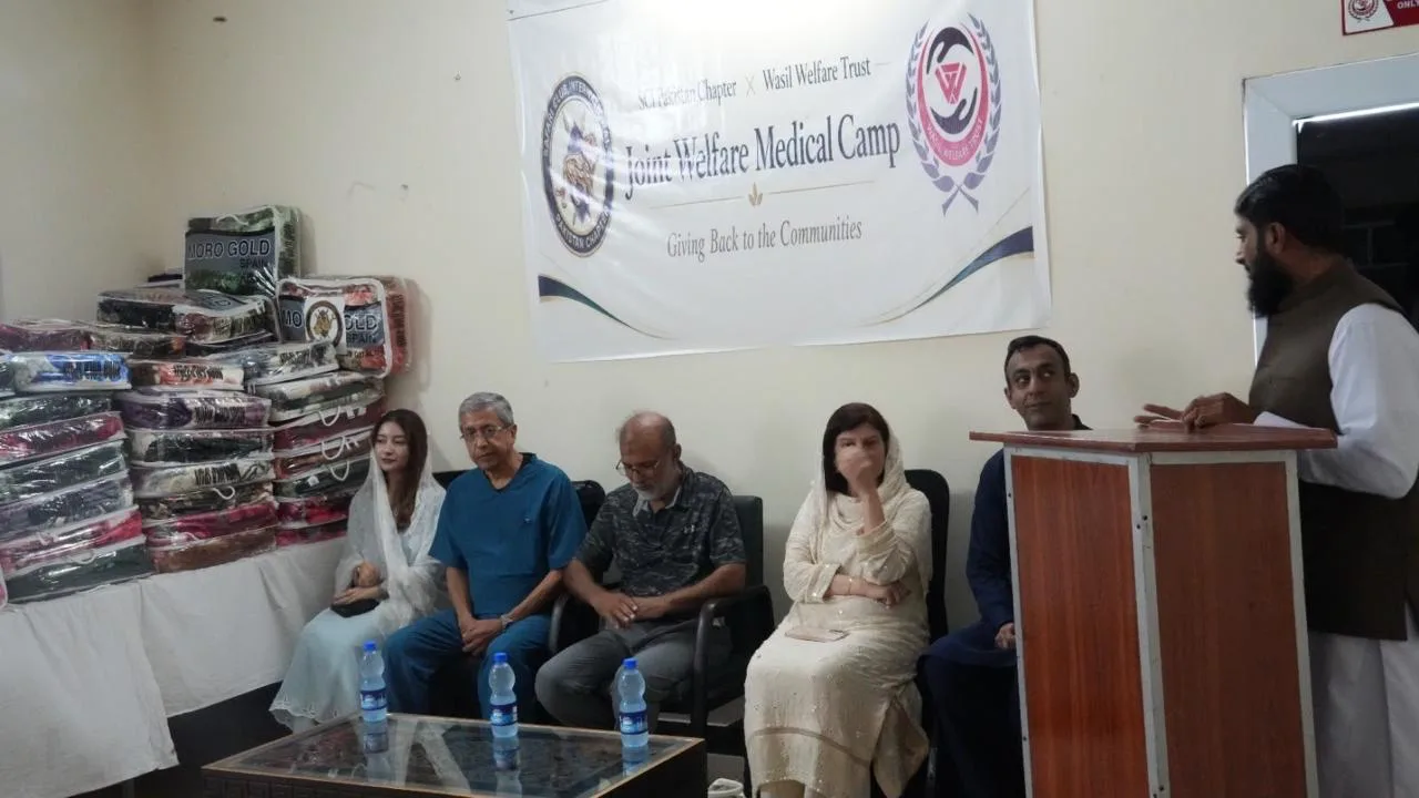 Guests sit beside stacked winter blankets under the Joint Welfare Medical Camp banner as a speaker addresses them.