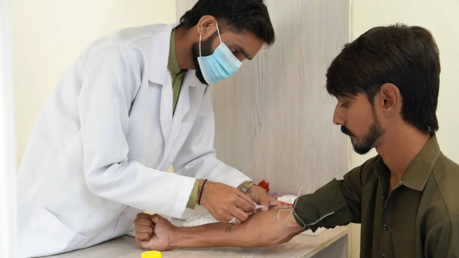 A doctor in a white coat and mask draws blood from a patient’s arm.