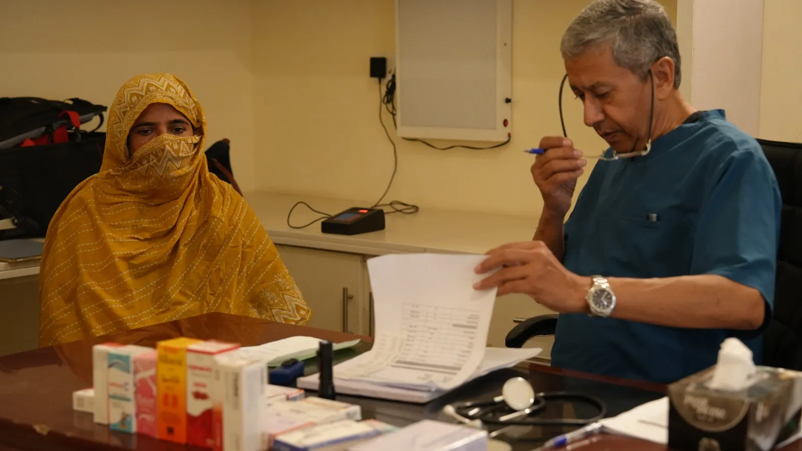 A doctor in blue scrubs reviews notes with a patient in a yellow headscarf.
