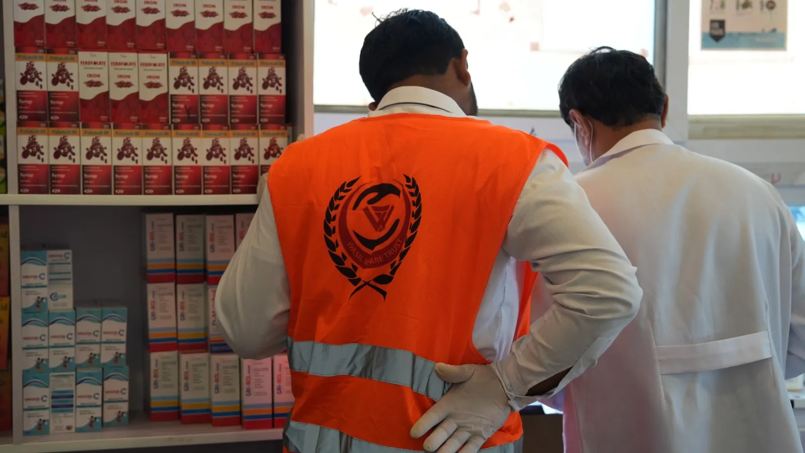 A volunteer in an orange vest stands before pharmacy shelves of boxed medicines.