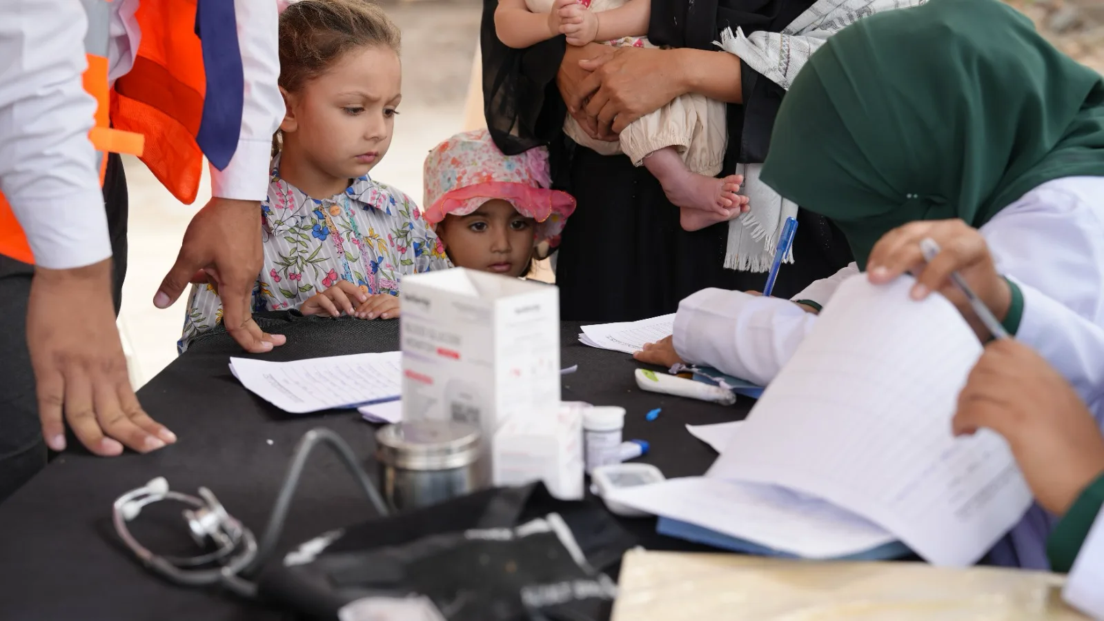 Two young girls watch as a doctor in a green headscarf fills in a registration sheet at the medical camp.