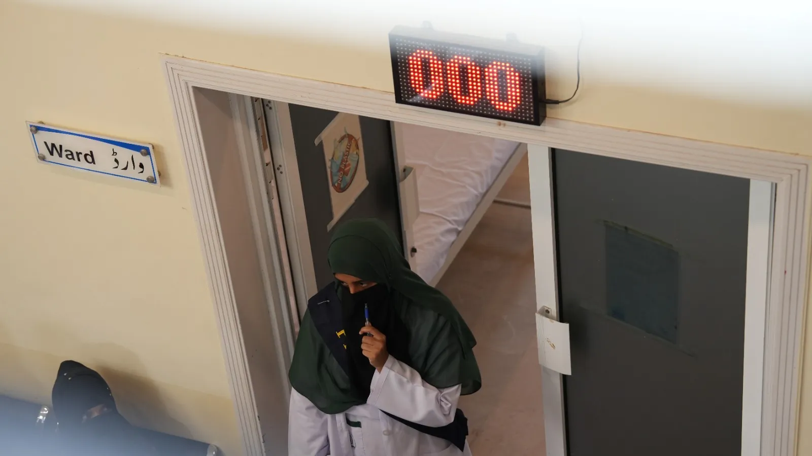 A woman in a headscarf walks through a ward doorway beneath a number display.