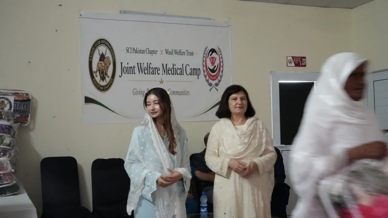 Two women in pale dress stand beneath the Joint Welfare Medical Camp banner as another woman walks past.