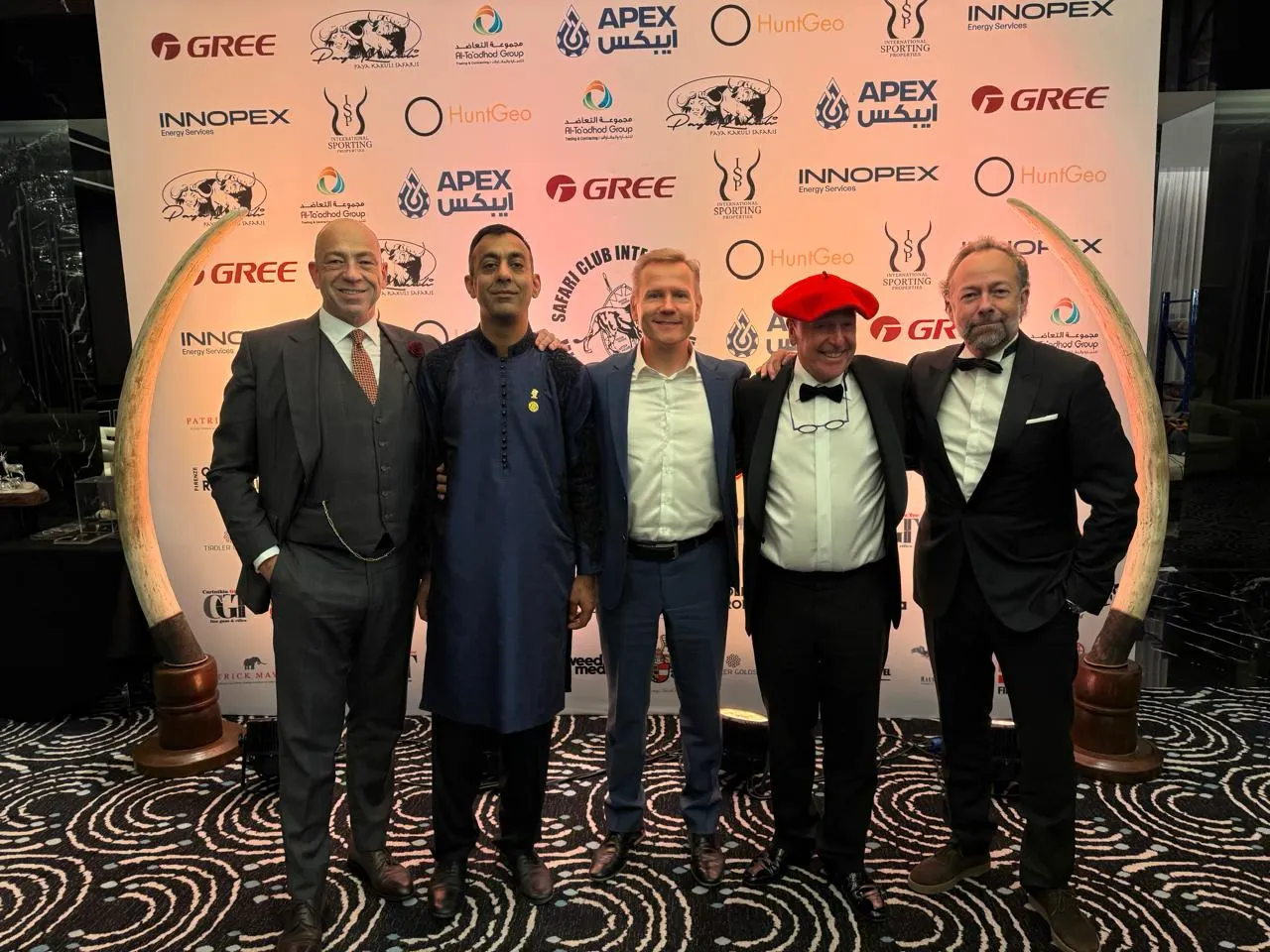 A chapter representative in a navy kurta stands arm in arm with four SCI members in front of a Safari Club International sponsor backdrop.