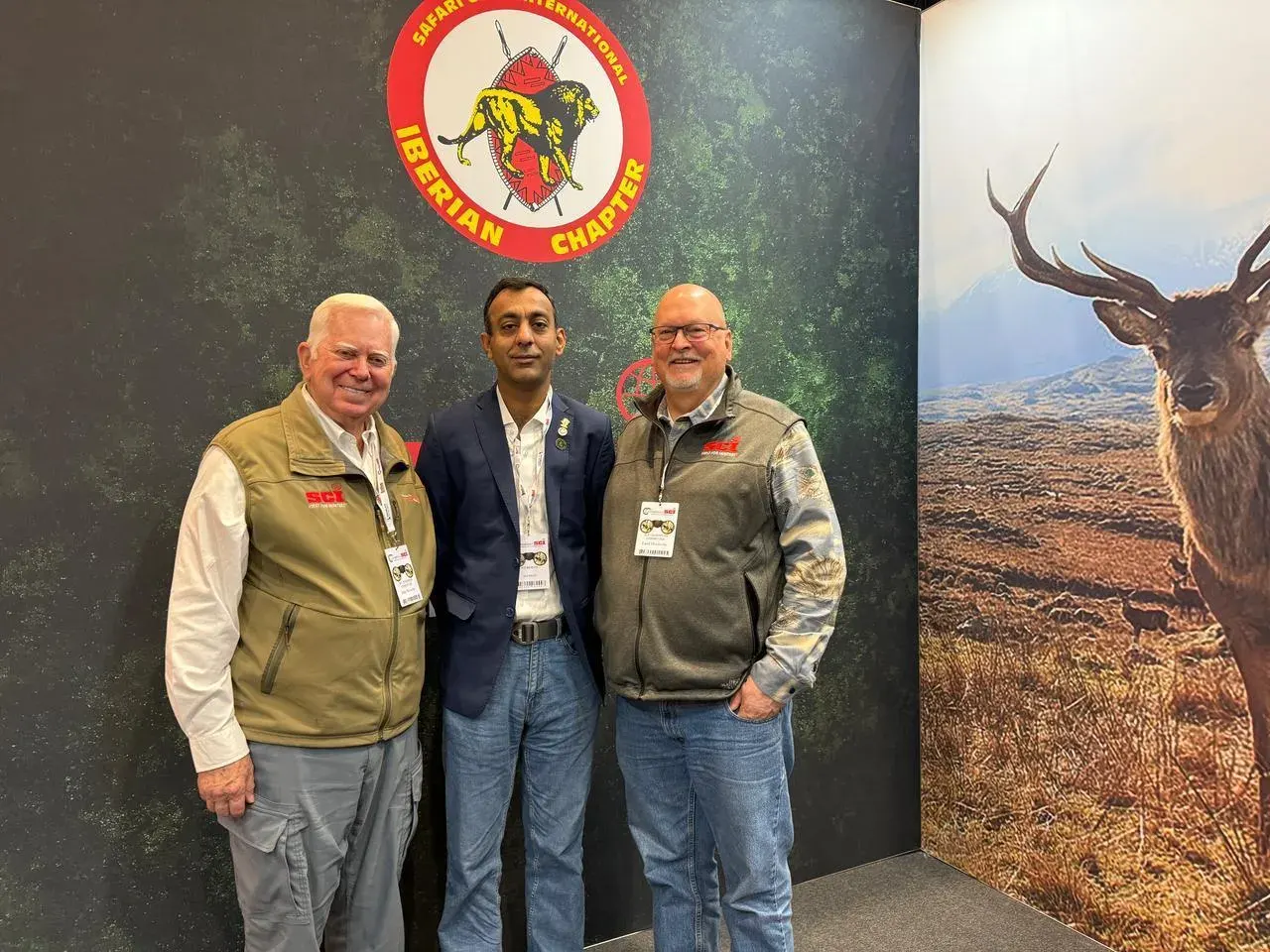 Three members stand together at the SCI Iberian Chapter stand, in front of a forest-green backdrop and a photograph of a red deer on open moorland.